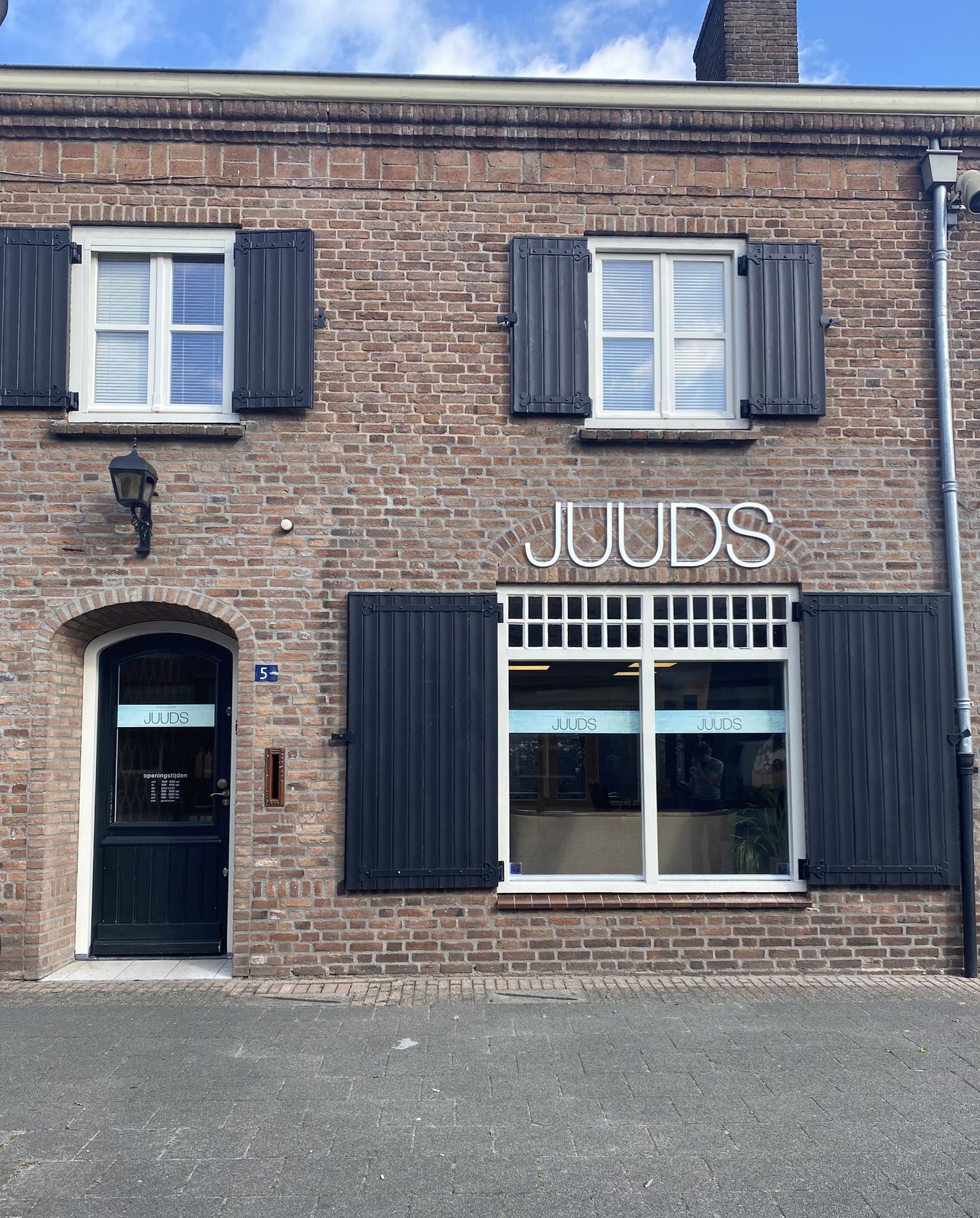 De bakstenen gevel van Kapsalon Juuds met de witte letters JUUDS boven het raam en zwarte luiken